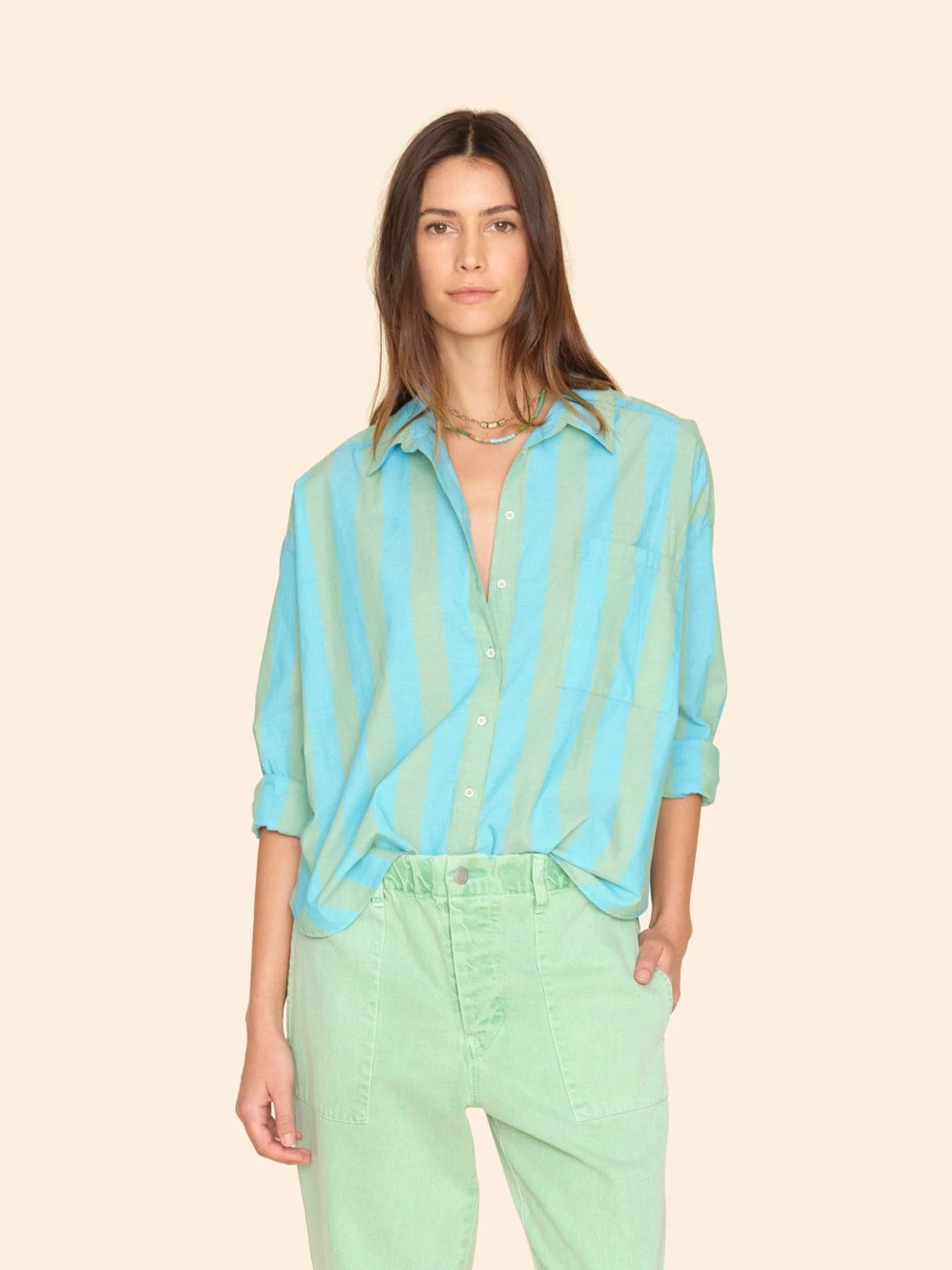 A person with long hair is wearing the Jordy Shirt by Xirena, a loose-fitting, button-front top featuring blue and green stripes with cuffed sleeves, paired with light green pants. They stand against a plain beige background.