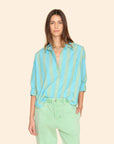 A person with long hair is wearing the Jordy Shirt by Xirena, a loose-fitting, button-front top featuring blue and green stripes with cuffed sleeves, paired with light green pants. They stand against a plain beige background.