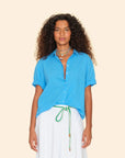 A person with curly hair sports a Xirena Channing Shirt in woven cotton poplin, a white skirt with a green and white rope belt, and several necklaces. They stand against a light beige background, effortlessly embodying the Los Angeles design aesthetic.