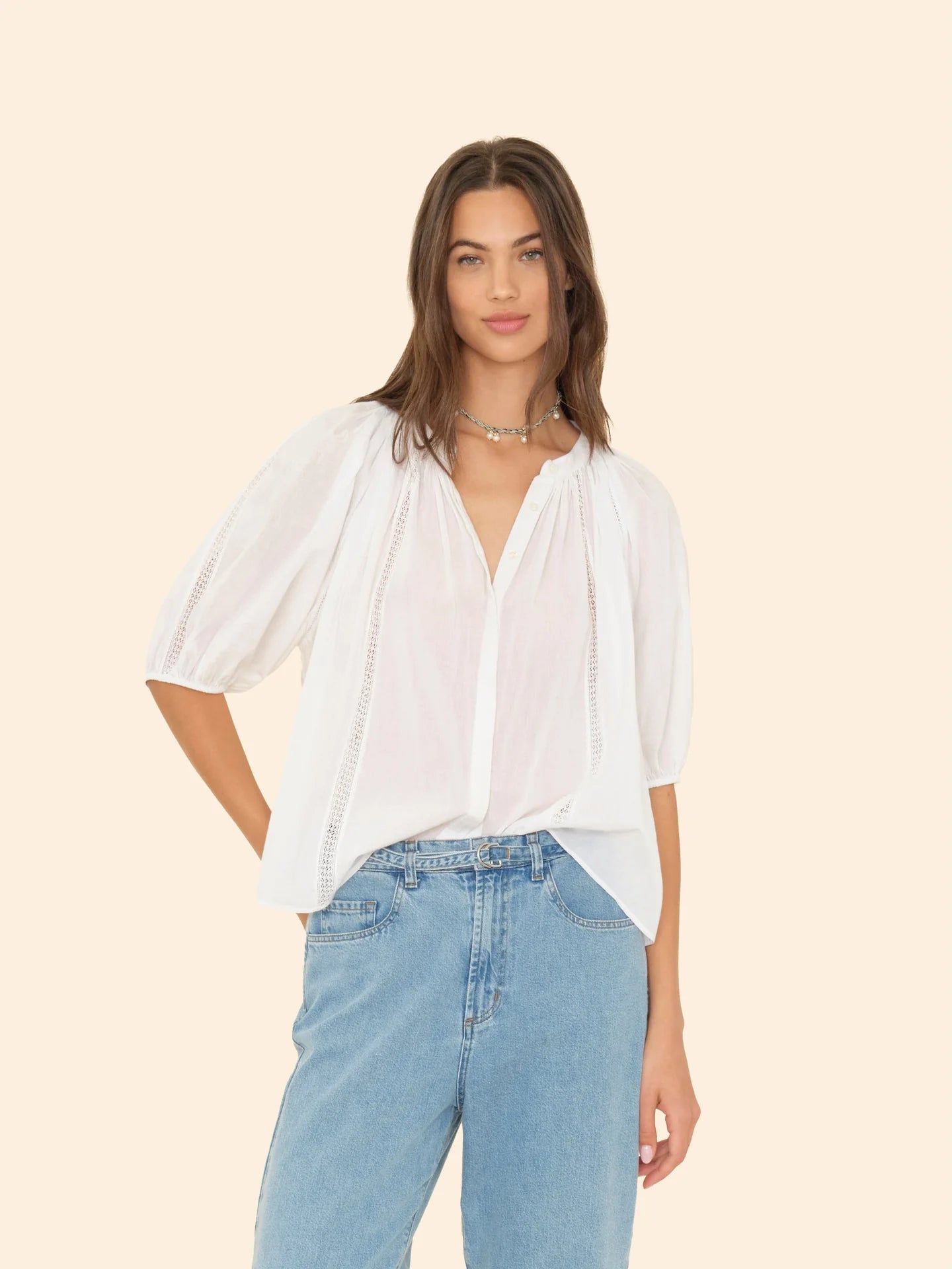 A person with long hair wearing the Evynn Shirt by Xirena, a white shirred blouse crafted from superfine cotton voile, along with blue jeans, stands against a plain background. The shirt features loose sleeves adorned with delicate lace insets.