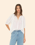 A person with long hair wearing the Evynn Shirt by Xirena, a white shirred blouse crafted from superfine cotton voile, along with blue jeans, stands against a plain background. The shirt features loose sleeves adorned with delicate lace insets.