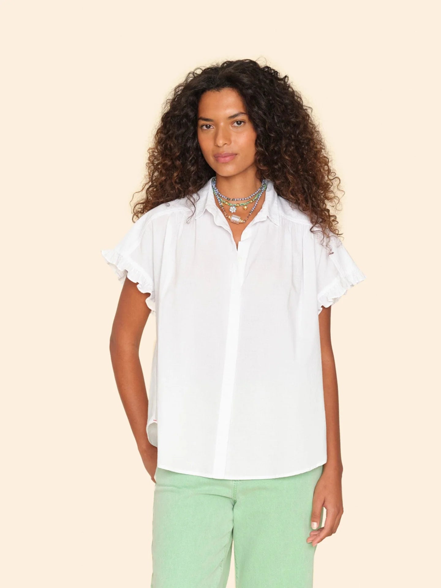 A person with curly hair is wearing the Noah Shirt by Xirena, a white cotton poplin blouse with ruffle sleeves, paired with green pants against a beige background. They are accessorized with layered necklaces.