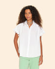 A person with curly hair is wearing the Noah Shirt by Xirena, a white cotton poplin blouse with ruffle sleeves, paired with green pants against a beige background. They are accessorized with layered necklaces.