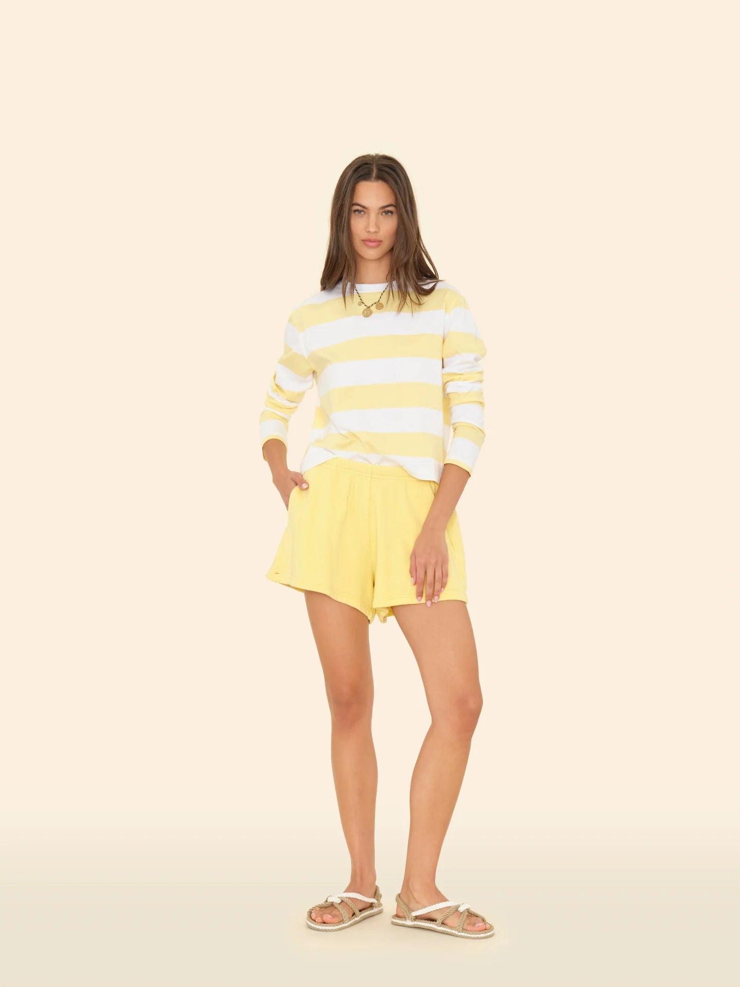 A woman stands against a light background wearing a yellow and white striped long-sleeve shirt, Shayne Sweatshorts by Xirena, beige sandals, and maintains a neutral expression with relaxed hands at her sides.