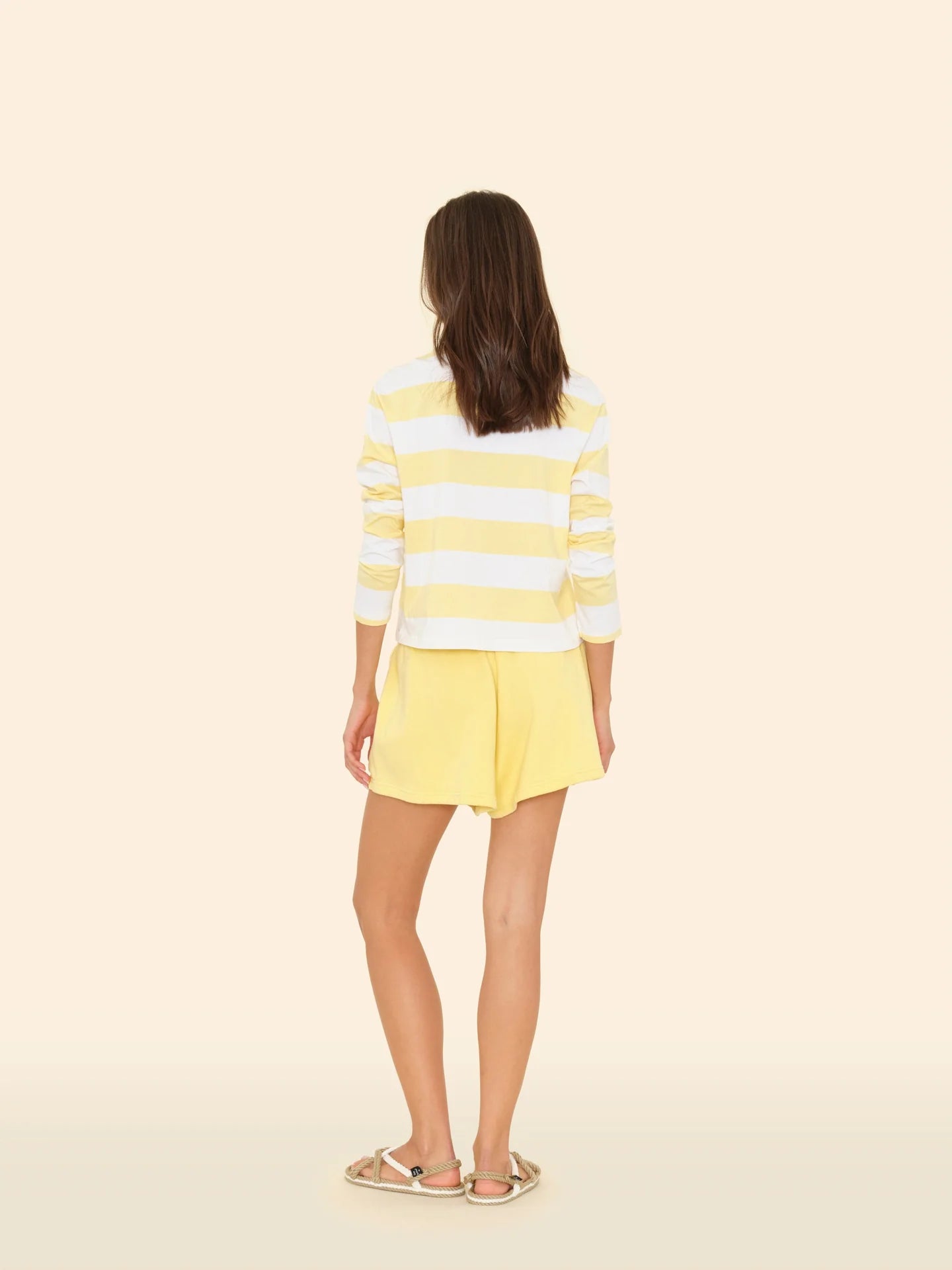 A person with long hair stands facing away, wearing a yellow and white striped long-sleeve shirt and the cozy Shayne Sweatshort by Xirena. They sport sandals against a plain beige background.