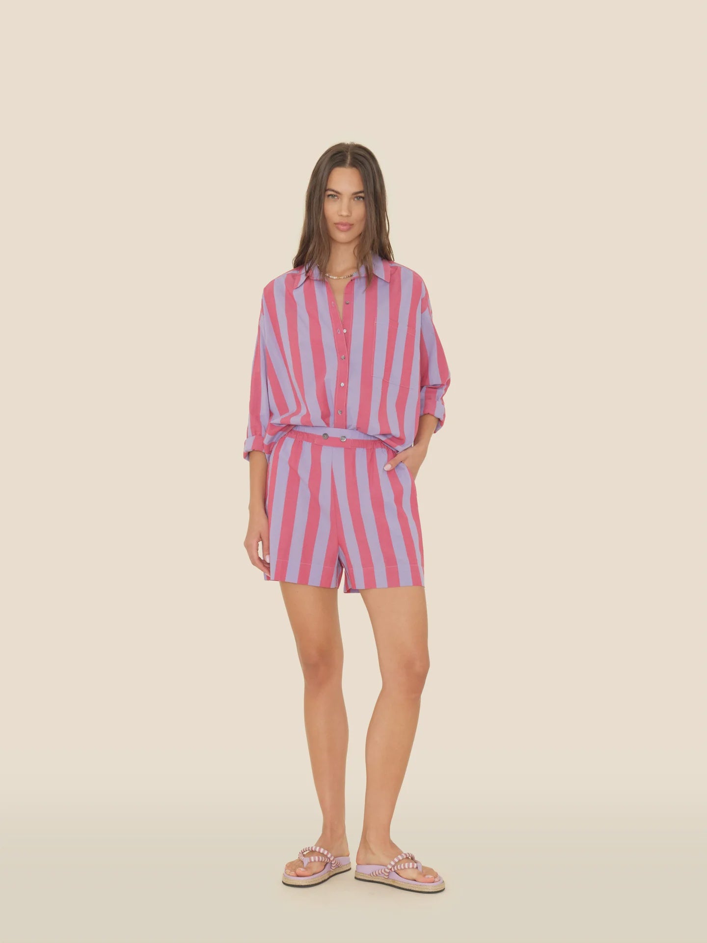 A person wearing an Amalfi Candy Stripe ensemble stands against a neutral background, featuring a pink and purple-striped button-up shirt tucked into Xirena’s Casey Shorts. Paired with flip-flops, their long hair perfectly complements the vibrant look.