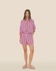 A person wearing an Amalfi Candy Stripe ensemble stands against a neutral background, featuring a pink and purple-striped button-up shirt tucked into Xirena’s Casey Shorts. Paired with flip-flops, their long hair perfectly complements the vibrant look.