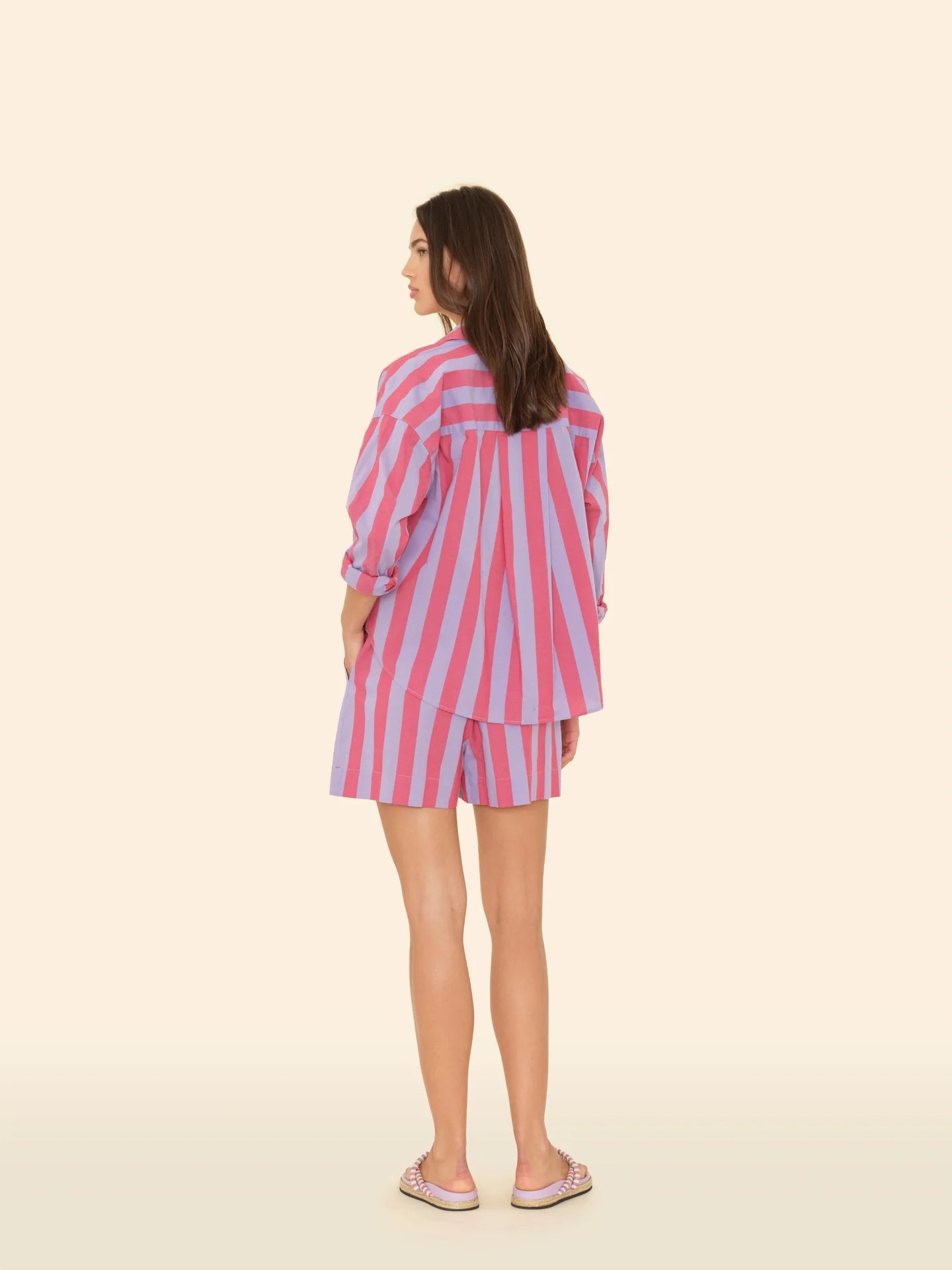 A woman stands facing away against a light background, dressed in an Amalfi Candy Stripe shirt paired with Xirena's Casey Short. She completes her look with playful pink shoes.