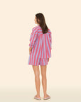 A woman stands facing away against a light background, dressed in an Amalfi Candy Stripe shirt paired with Xirena's Casey Short. She completes her look with playful pink shoes.