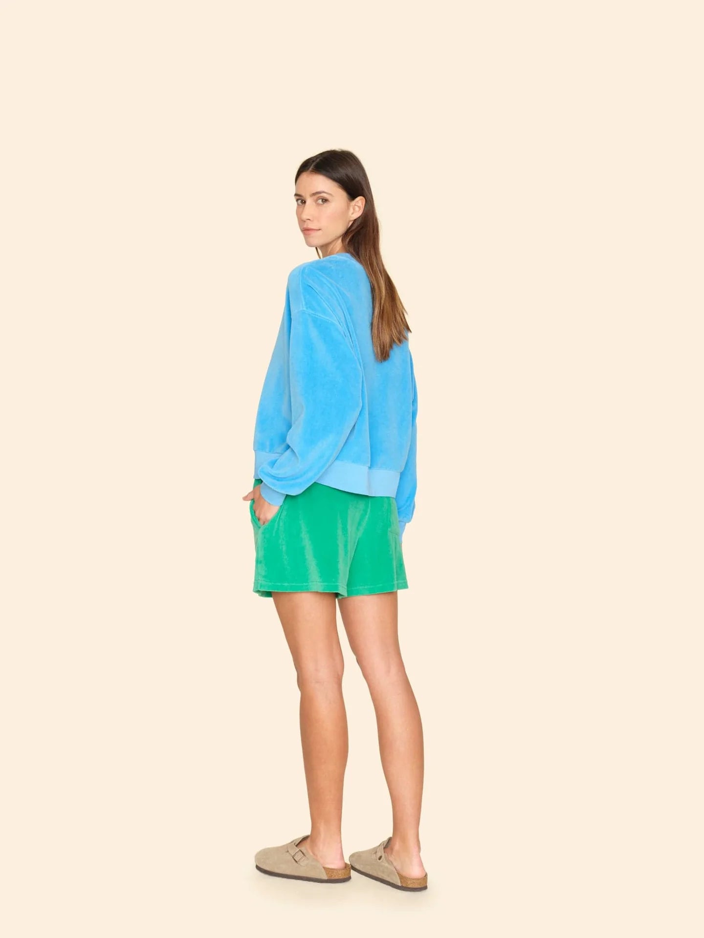 A person with long hair, clad in a blue sweater and Shayden Sweatshort by Xirena, featuring an elastic waist, looks over their shoulder. They wear beige sandals against a beige backdrop, embodying Los Angeles' relaxed vibe.