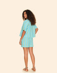 A person with curly hair stands with their back to the camera, dressed in a blue and turquoise outfit consisting of a loose shirt and Xirena's Casey Short. Barefoot against a simple beige background, they exude an effortless, laid-back vibe.