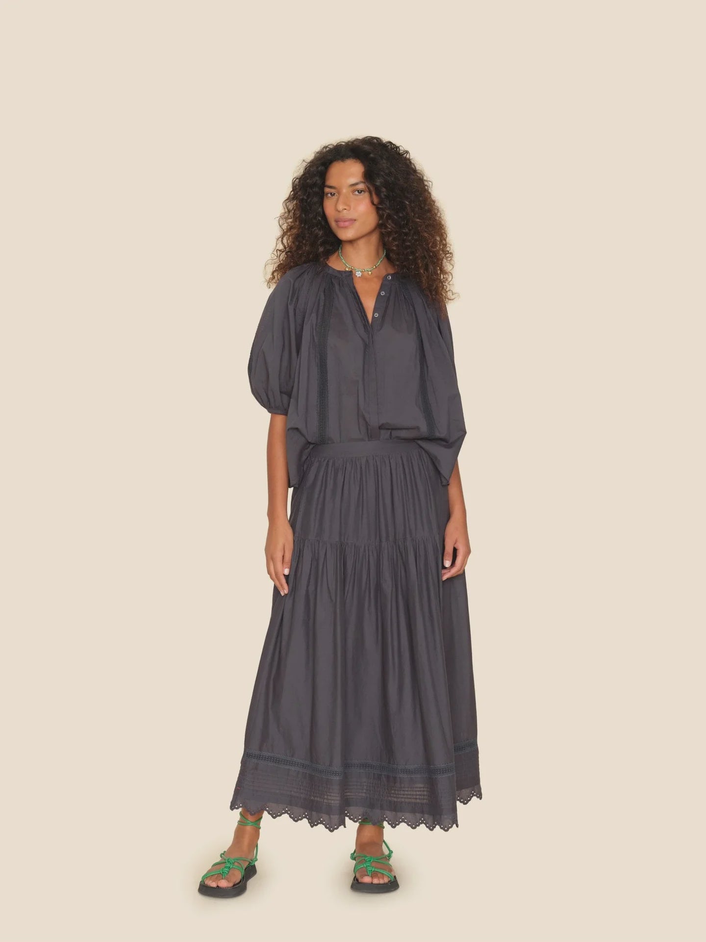 A woman with curly hair is wearing a dark gray, loose-fitting blouse combined with the Tilda Skirt from Xirena, crafted from cotton voile in an A-line silhouette. Her look is completed with green sandals and a necklace as she stands against a plain beige background.