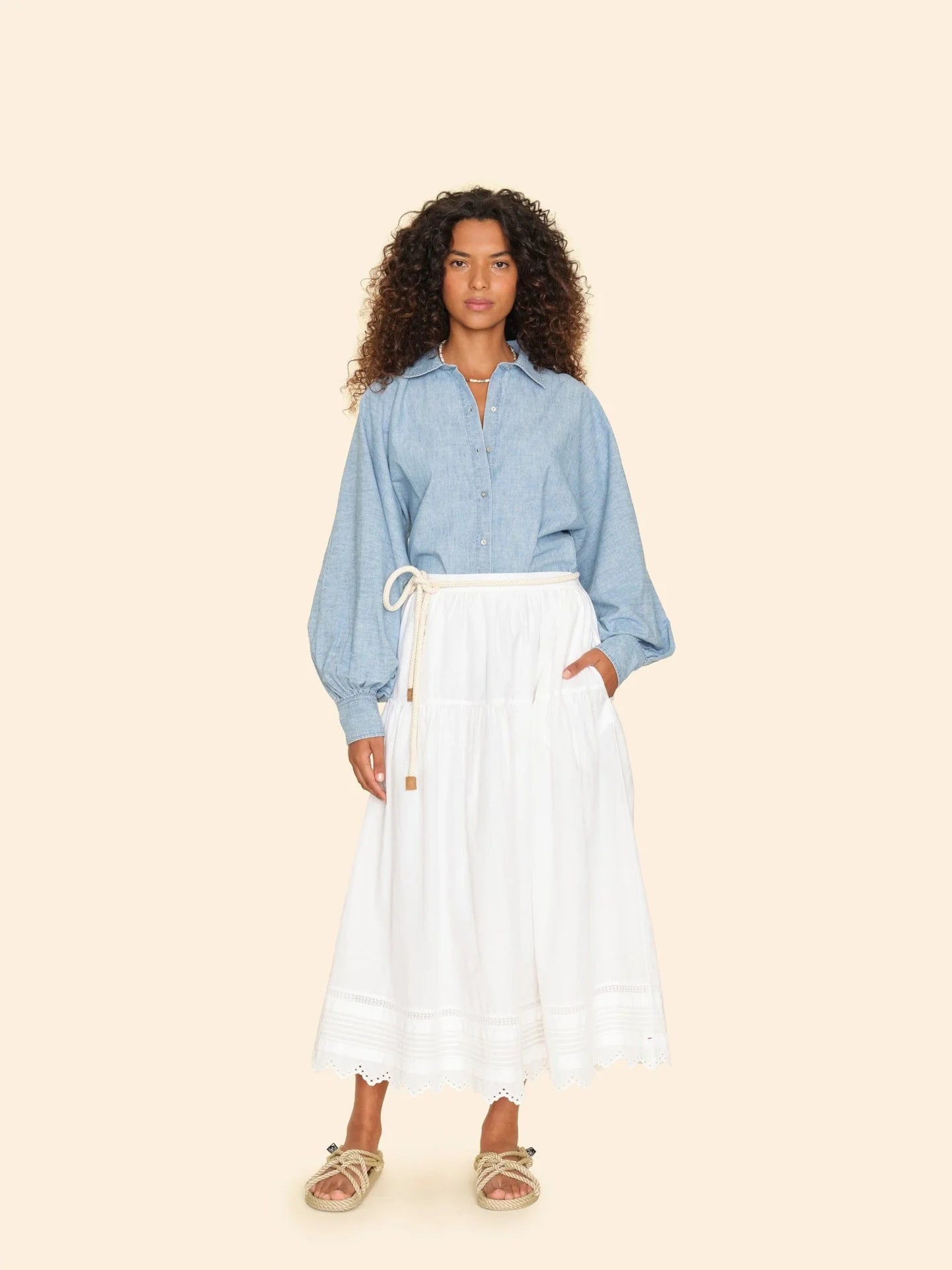 A person with curly hair stands against a beige background, wearing a light blue denim shirt and an elegant white Tilda Skirt by Xirena featuring a scalloped eyelet hem. They complete their look with tan espadrille sandals.