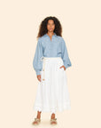 A person with curly hair stands against a beige background, wearing a light blue denim shirt and an elegant white Tilda Skirt by Xirena featuring a scalloped eyelet hem. They complete their look with tan espadrille sandals.