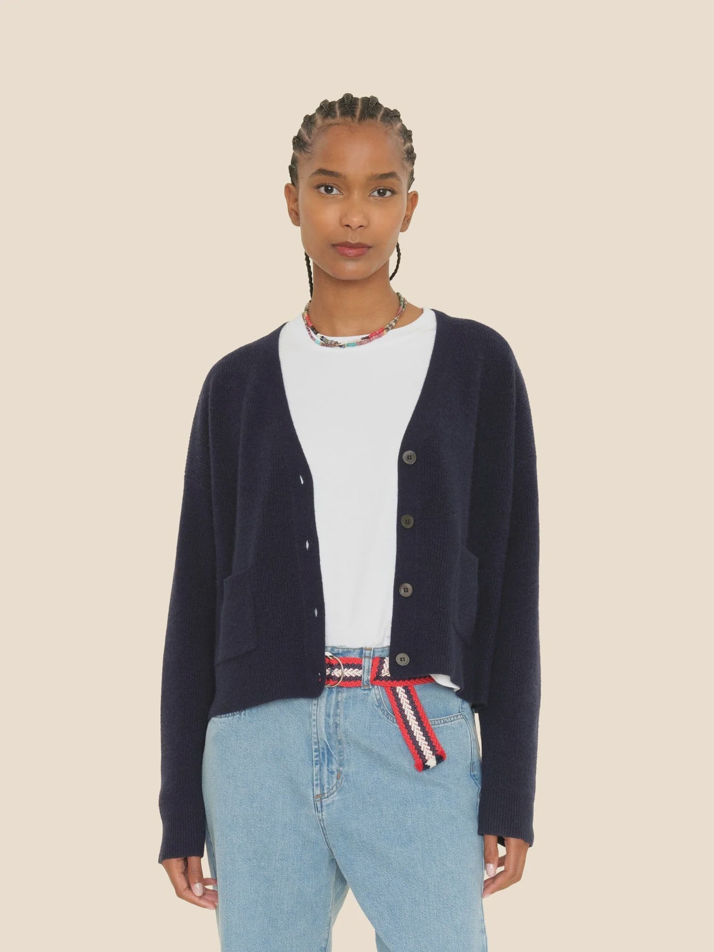 The individual with braided hair is dressed in a navy "Trudie Cardigan" by Xirena over a white T-shirt and light blue jeans, accessorized with a red, white, and blue belt, standing against a plain background.