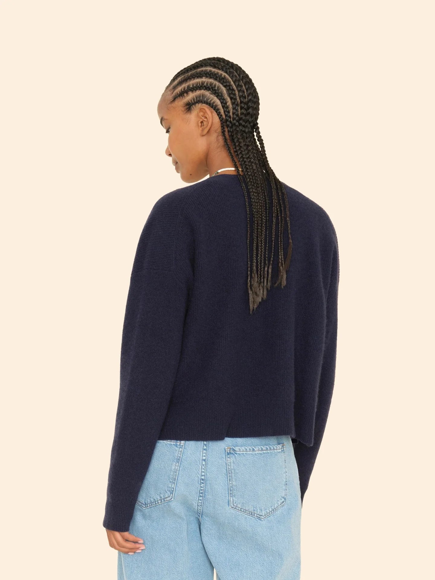 A person with long braided hair, wearing the Trudie Cardigan by Xirena paired with light blue jeans, is turned away against a plain beige background.