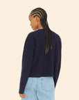 A person with long braided hair, wearing the Trudie Cardigan by Xirena paired with light blue jeans, is turned away against a plain beige background.