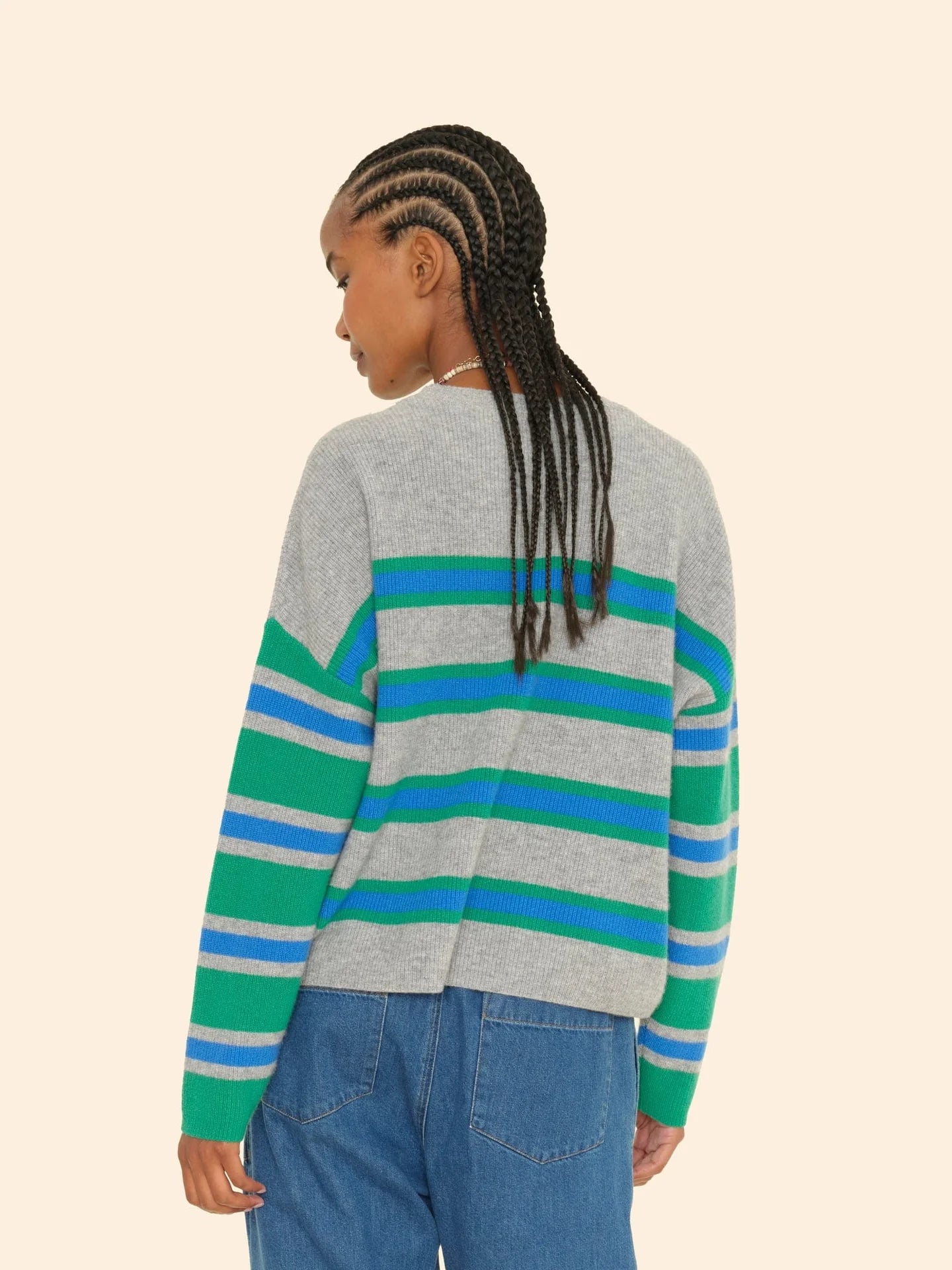 A person with braided hair is seen from the back, wearing a Lela Sweater by Xirena, a gray cashmere garment with blue and green stripes, accompanied by blue jeans. The background is a plain light beige color.