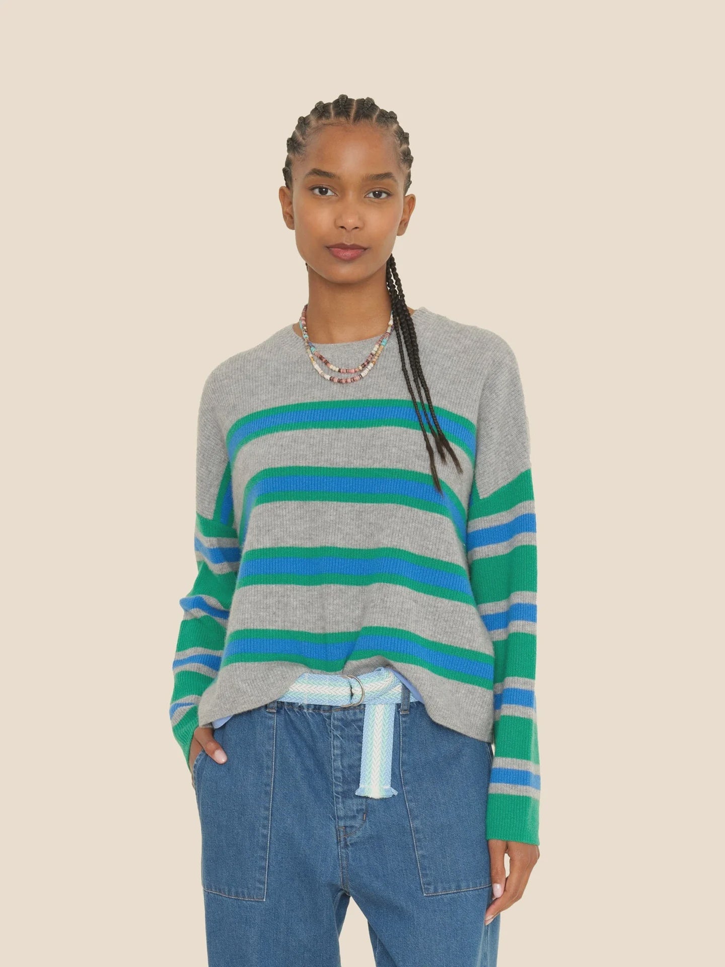 A person wearing the ultra-soft Xirena Lela Sweater, featuring vibrant blue and green stripes, stands against a beige background. They have long braided hair and are dressed in blue jeans paired with a light blue belt.