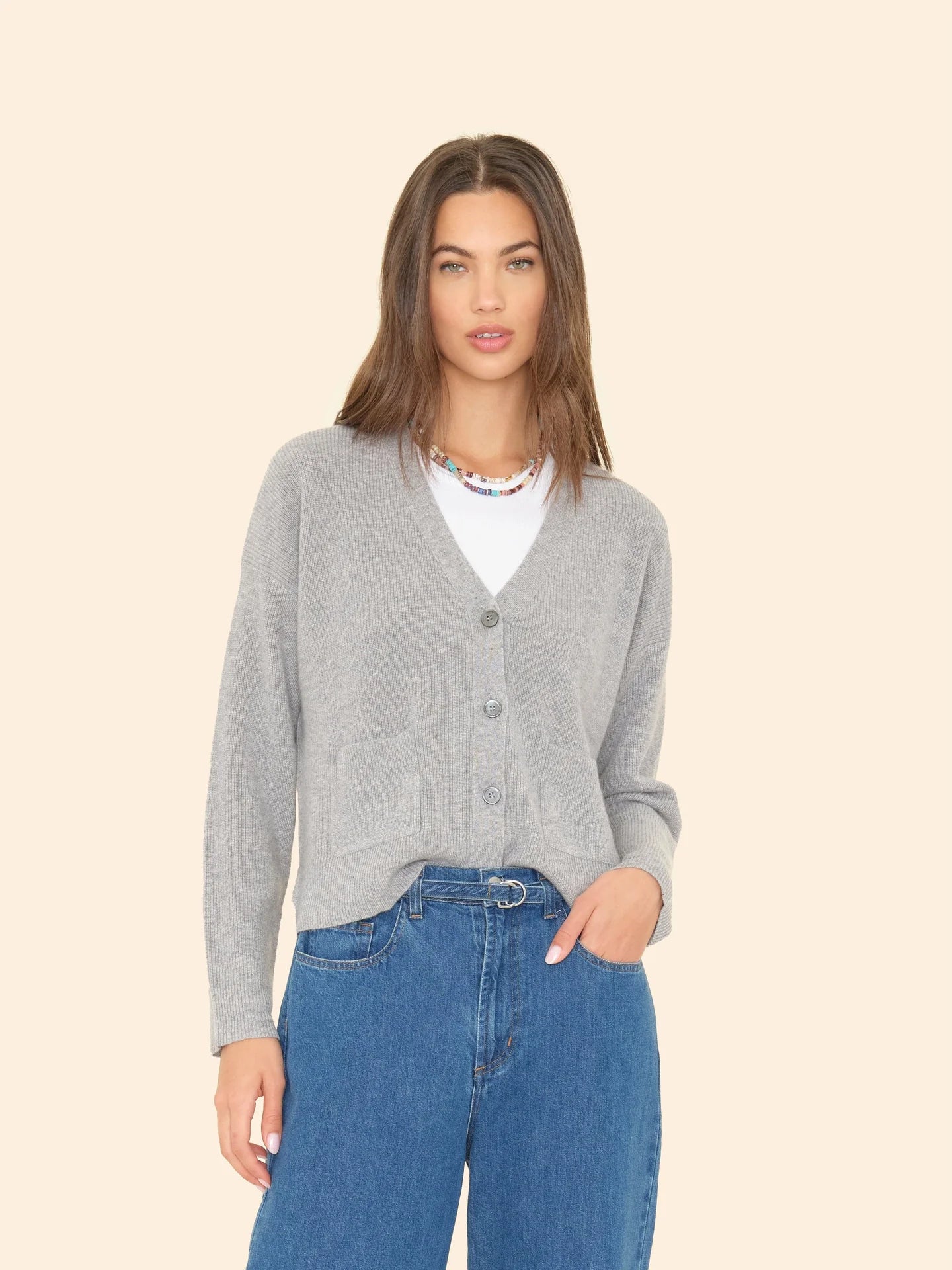 A person with long hair is dressed in Xirena's ultra-soft Trudie Cardigan in gray, layered over a white shirt and paired with blue jeans. They pose against a cream-colored background and are adorned with a vibrant beaded necklace.