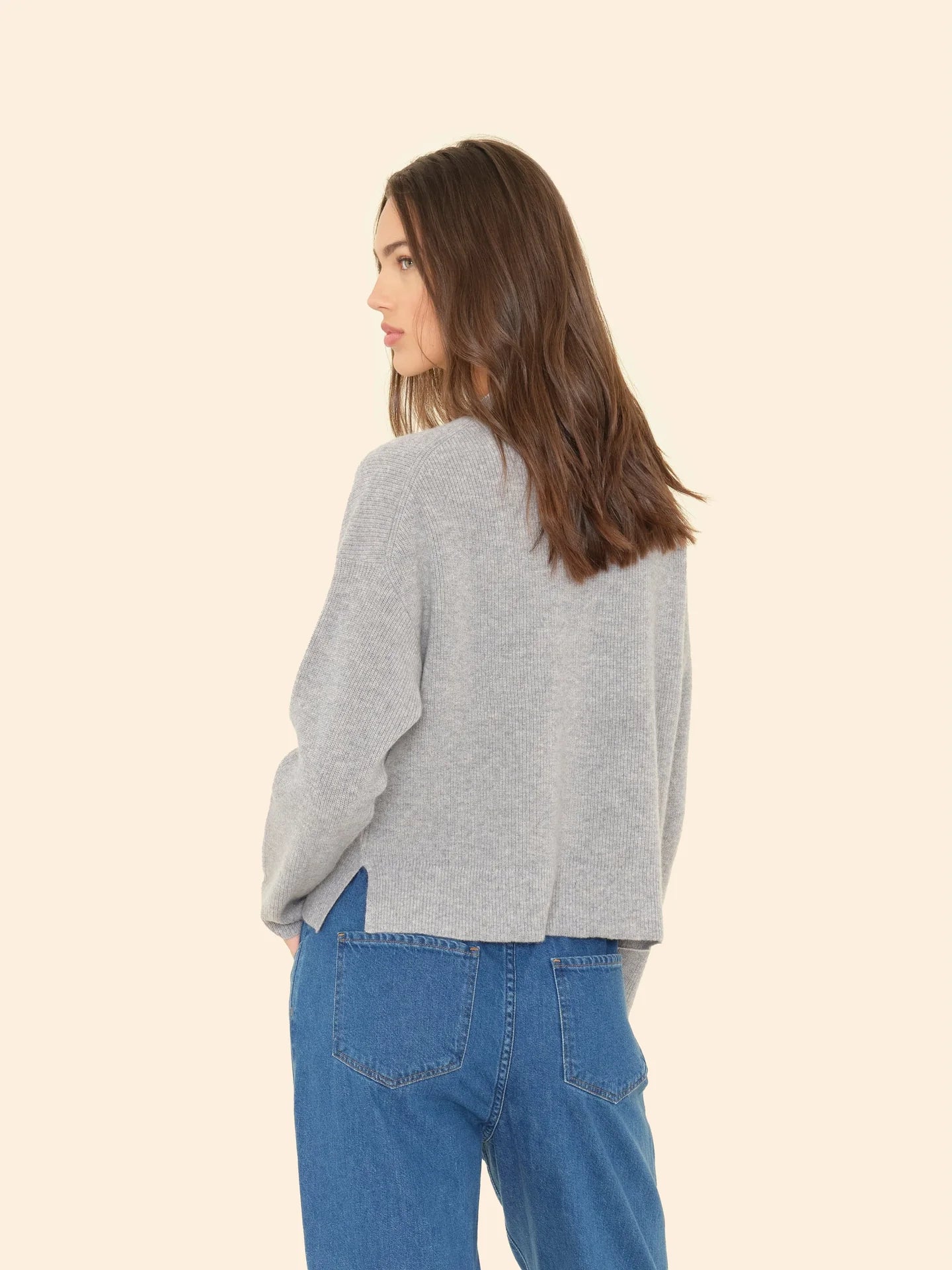 A person with long brown hair is turned slightly to the side, wearing the ultra-soft Trudie Cardigan by Xirena in gray and blue jeans. The background is a plain, light beige.