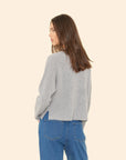 A person with long brown hair is turned slightly to the side, wearing the ultra-soft Trudie Cardigan by Xirena in gray and blue jeans. The background is a plain, light beige.