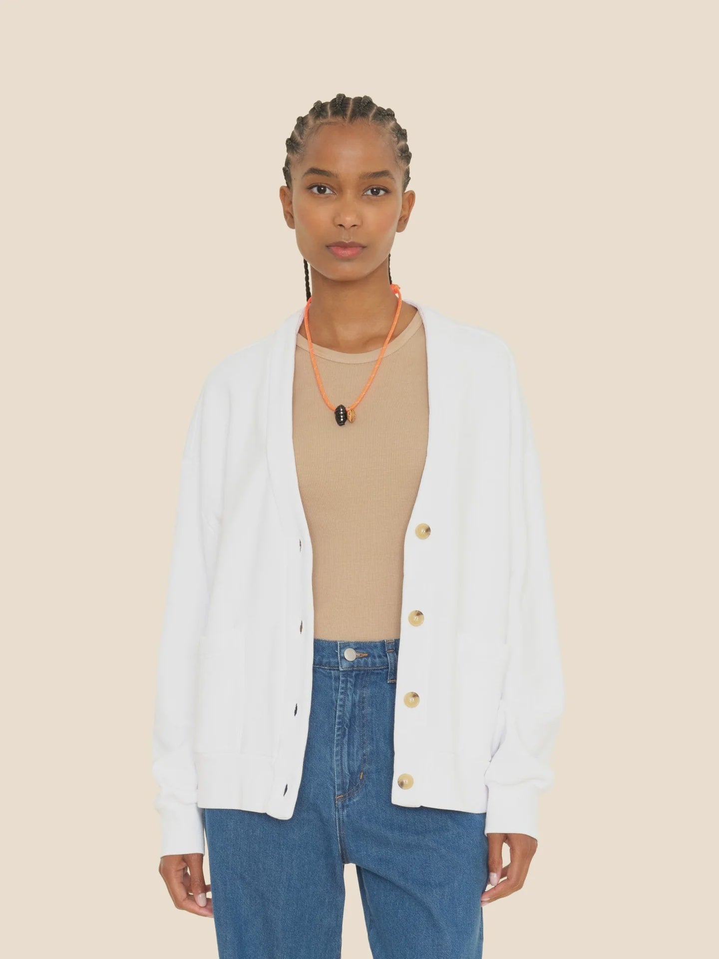 A person wearing the ultra-soft Benny Cardigan by Xirena in white, paired with a beige top and blue jeans, accessorized with an orange lanyard and styled braided hair, stands against a plain background.