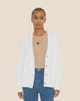 A person wearing the ultra-soft Benny Cardigan by Xirena in white, paired with a beige top and blue jeans, accessorized with an orange lanyard and styled braided hair, stands against a plain background.