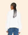 A person with long braided hair, wearing the Benny Cardigan from Xirena styled with jeans, is shown from behind against a beige background.