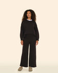 A person with curly hair stands against a beige background, wearing a black sweater and ultra-soft Xirena Emmette Sweatpants featuring wide legs and an elasticized waist, paired with beige shoes. They look confidently ahead.