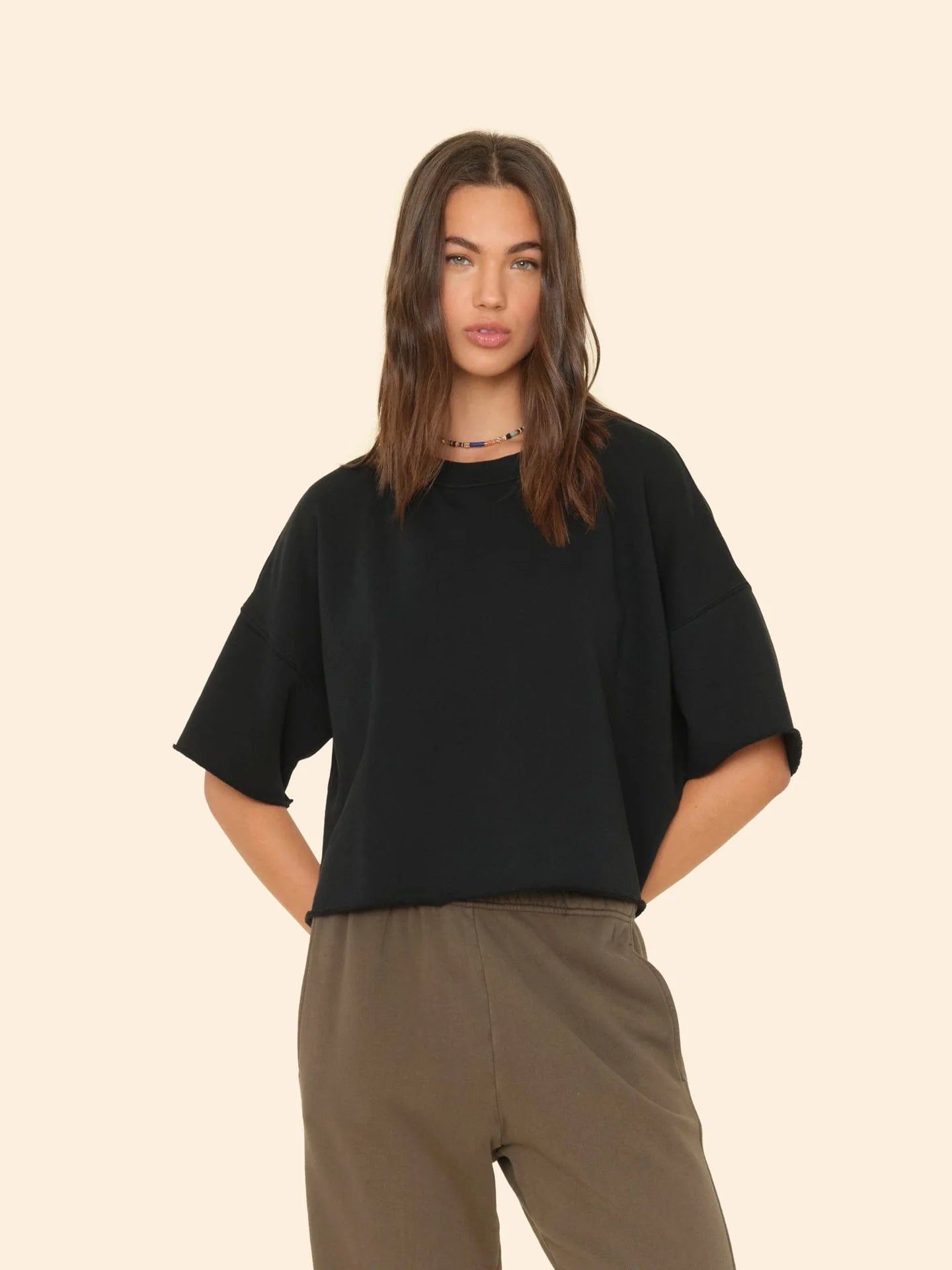 A person with long hair is wearing a black Romeo Sweatshirt by Xirena and khaki pants. They have their hands in their pockets, exuding elevated comfort as they stand against a plain beige background.