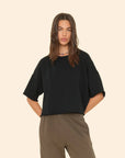 A person with long hair is wearing a black Romeo Sweatshirt by Xirena and khaki pants. They have their hands in their pockets, exuding elevated comfort as they stand against a plain beige background.