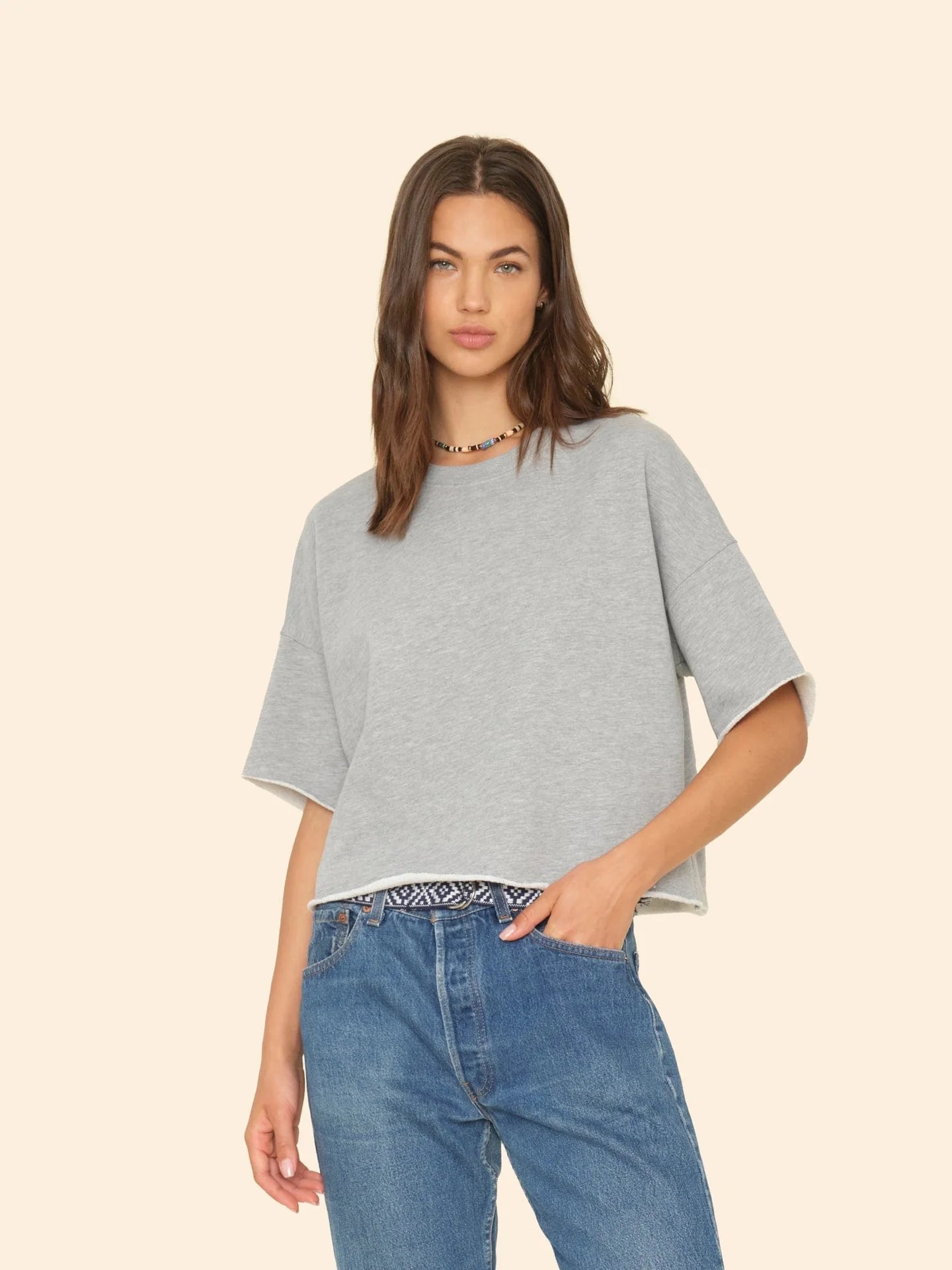 A person with long hair is wearing the Romeo Sweatshirt by Xirena, featuring raw edge sleeves, paired with blue jeans. They stand against a beige background with a neutral expression and are adorned with a beaded necklace.