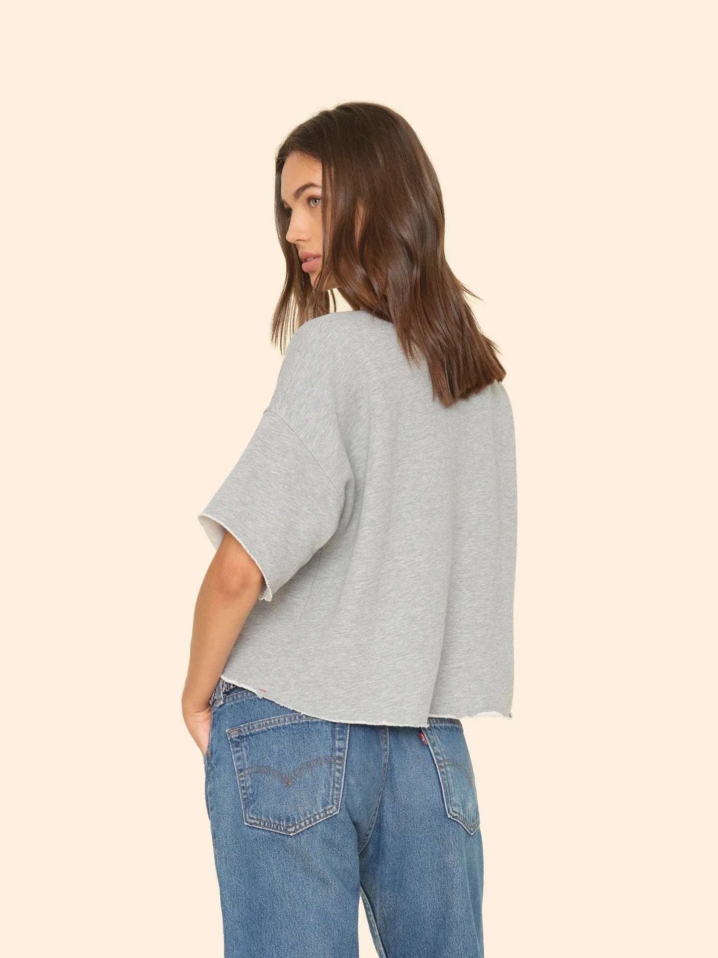 A woman with shoulder-length brown hair is wearing the Romeo Sweatshirt by Xirena, featuring raw edge sleeves, paired with blue jeans. She stands partially turned, looking over her shoulder against a plain beige background, embodying elevated comfort.