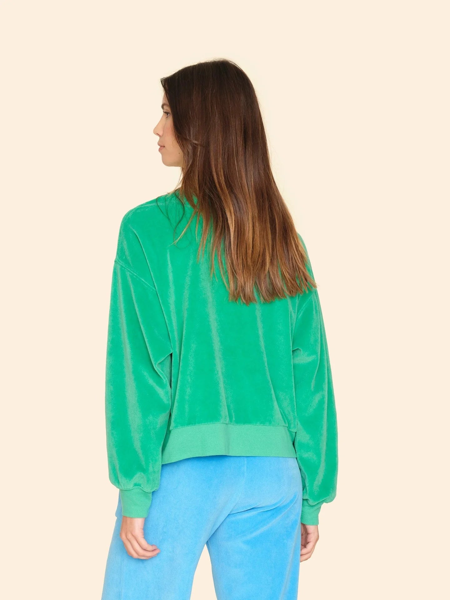 A person with long brown hair is standing with their back to the camera, dressed in the vintage-inspired Huxley Sweatshirt by Xirena in green velour and blue pants against a beige background.