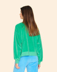 A person with long brown hair is standing with their back to the camera, dressed in the vintage-inspired Huxley Sweatshirt by Xirena in green velour and blue pants against a beige background.