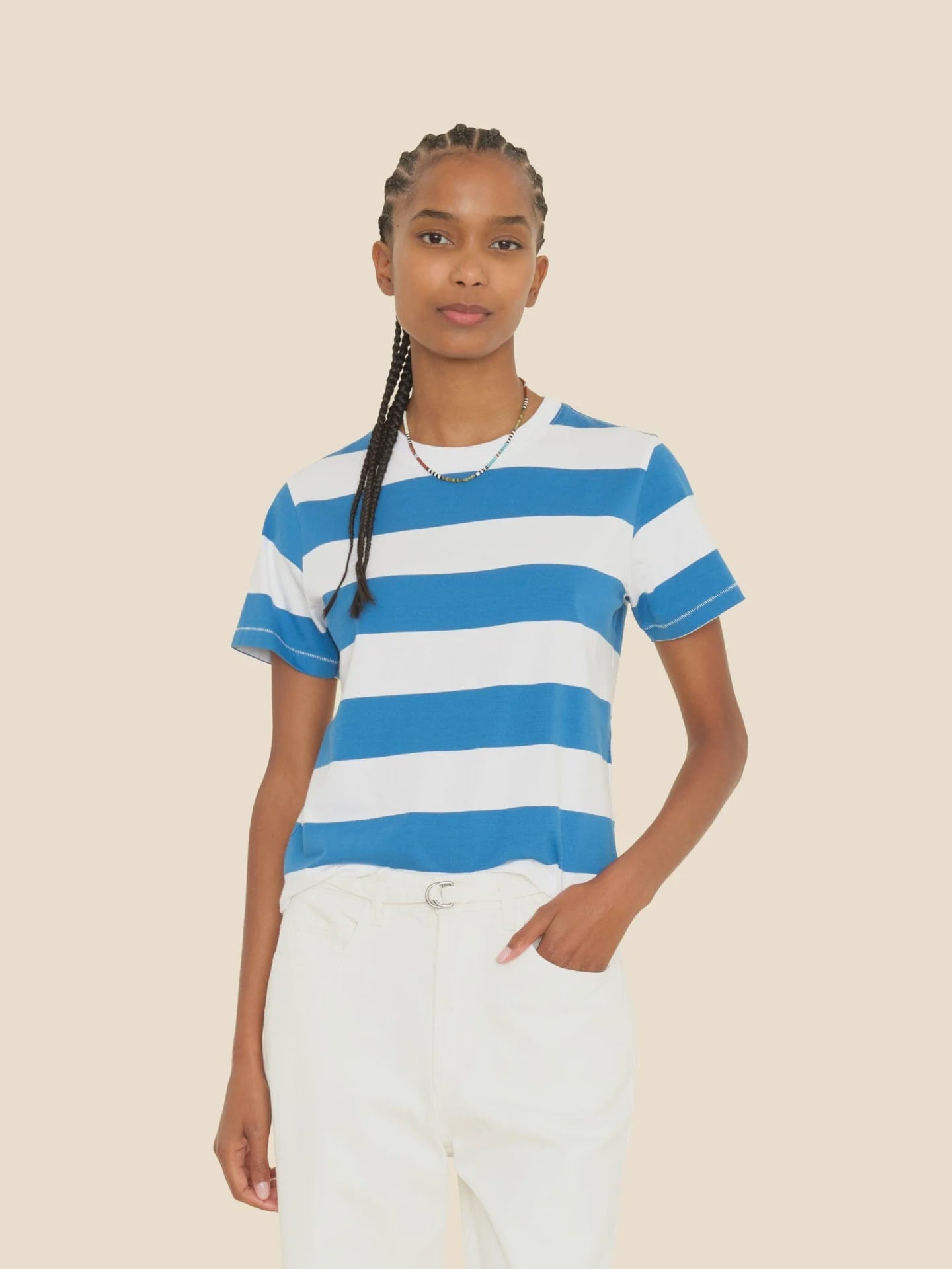 A person with braided hair wears the Xirena Georgy Tee, a vintage-inspired design featuring blue and white stripes, paired with white pants. Standing against a plain beige background, the boxy fit of the striped cotton jersey tee complements their casual stance as they place one hand in their pocket and maintain a neutral expression.