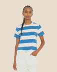 A person with braided hair wears the Xirena Georgy Tee, a vintage-inspired design featuring blue and white stripes, paired with white pants. Standing against a plain beige background, the boxy fit of the striped cotton jersey tee complements their casual stance as they place one hand in their pocket and maintain a neutral expression.