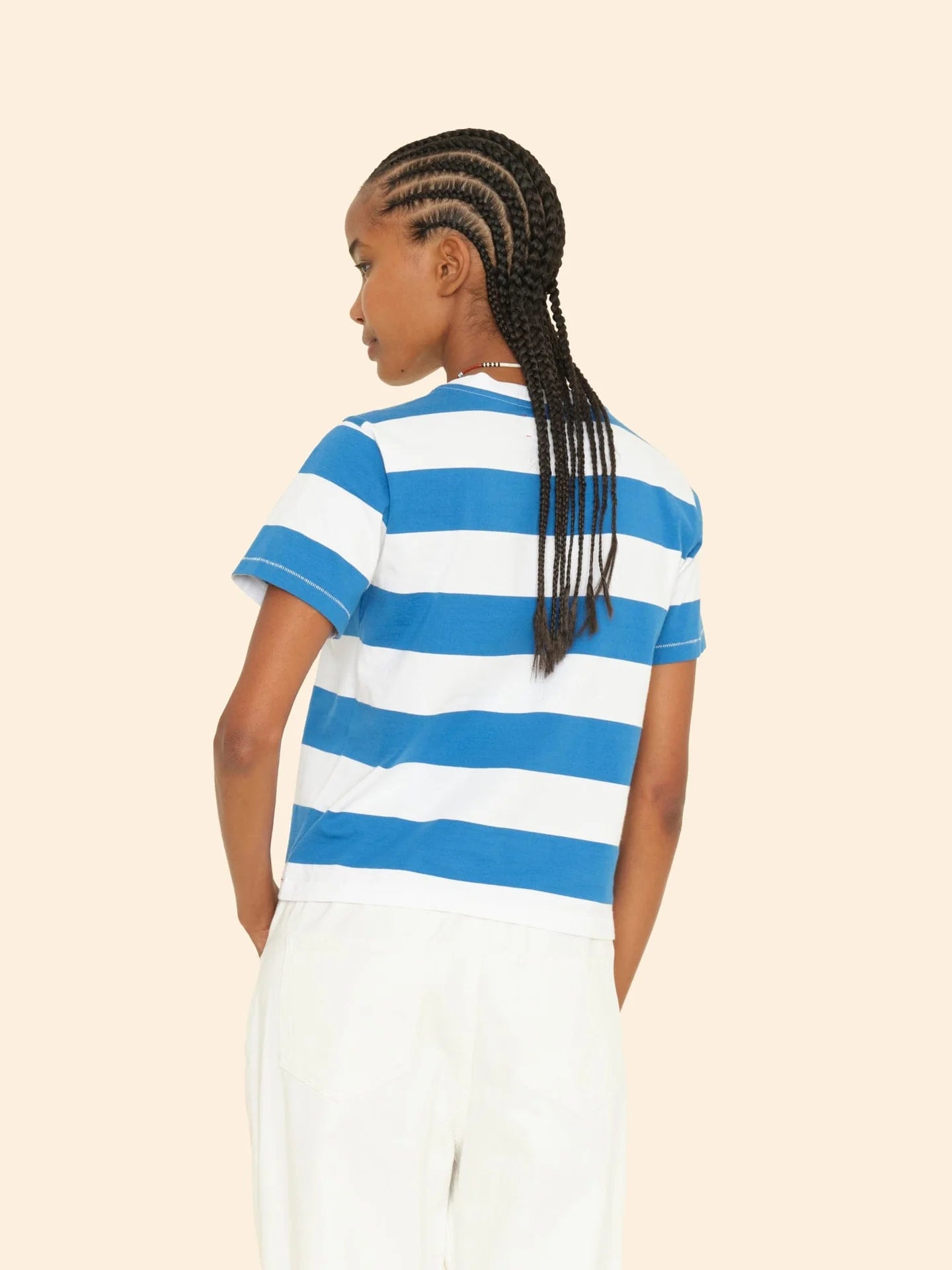 A person with braided hair is seen from behind wearing the Georgy Tee by Xirena, a vintage-inspired shirt in blue and white striped cotton jersey, paired with white pants. They are standing against a neutral background.