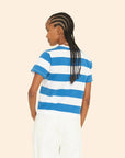 A person with braided hair is seen from behind wearing the Georgy Tee by Xirena, a vintage-inspired shirt in blue and white striped cotton jersey, paired with white pants. They are standing against a neutral background.