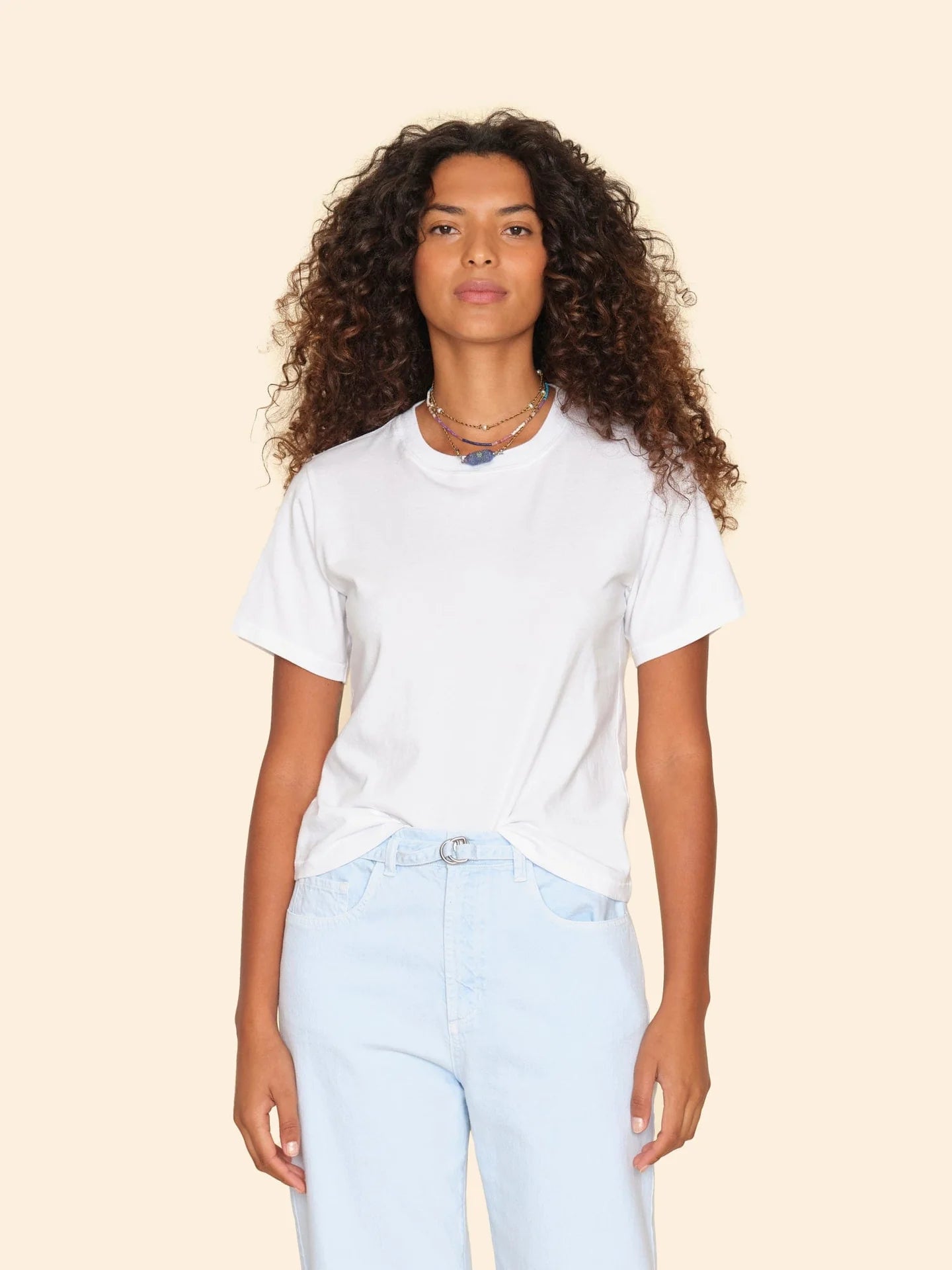 A person with long curly hair, wearing the Georgy Tee from Xirena and light blue jeans, stands against a light beige background. They have a neutral expression and are accessorized with layered necklaces.