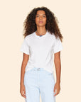 A person with long curly hair, wearing the Georgy Tee from Xirena and light blue jeans, stands against a light beige background. They have a neutral expression and are accessorized with layered necklaces.