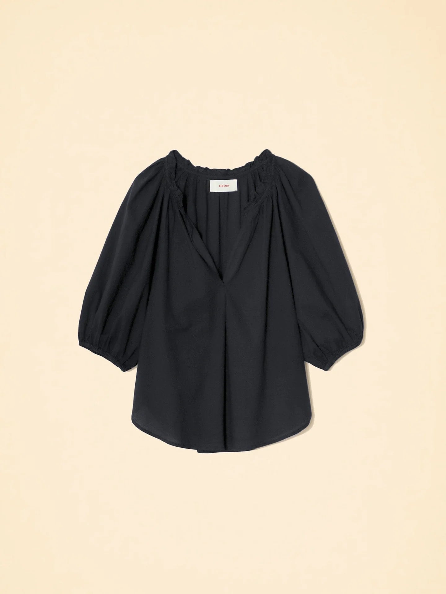 The Jules Top by Xirena is a black, loose-fitting cotton poplin blouse with three-quarter bubble sleeves and a gathered, elastic neckline, shown against a beige background.