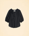 The Jules Top by Xirena is a black, loose-fitting cotton poplin blouse with three-quarter bubble sleeves and a gathered, elastic neckline, shown against a beige background.