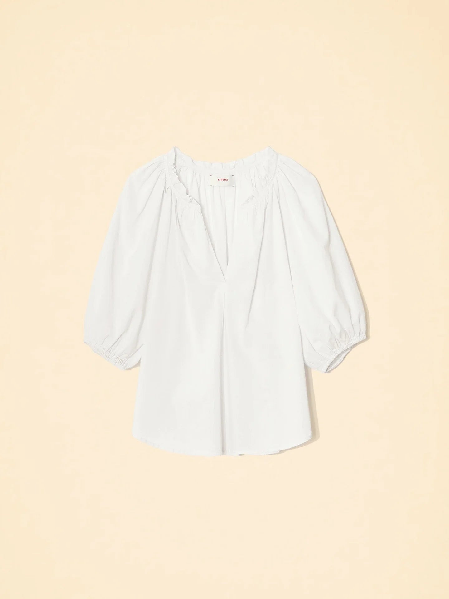 The Jules Top by Xirena is a white cotton poplin blouse featuring a loose fit, three-quarter bubble sleeves, a gathered neckline, and a V-shaped front slit, shown against a light beige background.