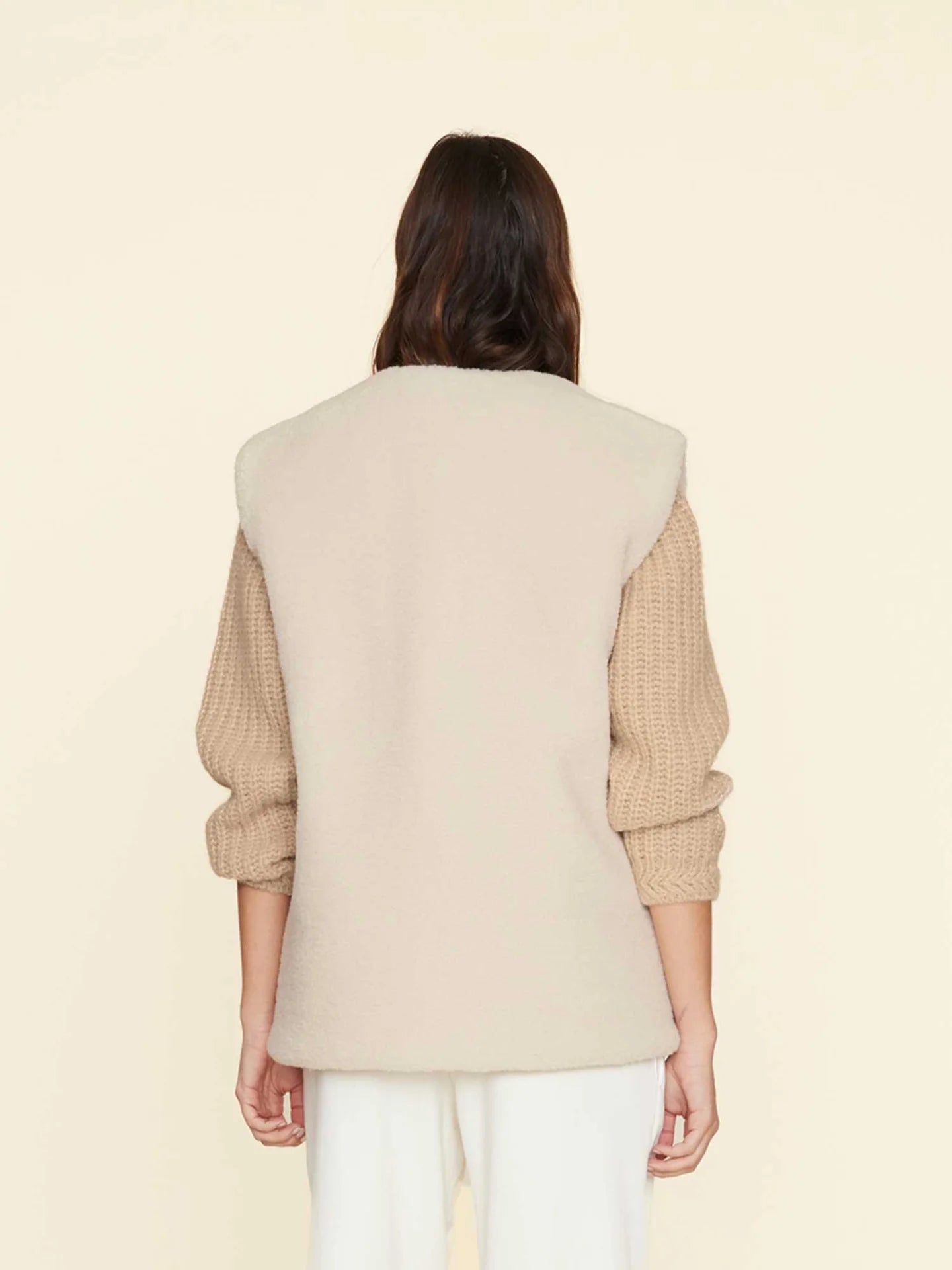 A person with long brown hair stands facing away, wearing the Thea Vest by Xirena, a cream-colored, sleeveless design featuring side slit pockets, over a beige knit sweater and white pants. The background is a soft pastel yellow.