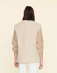 A person with long brown hair stands facing away, wearing the Thea Vest by Xirena, a cream-colored, sleeveless design featuring side slit pockets, over a beige knit sweater and white pants. The background is a soft pastel yellow.