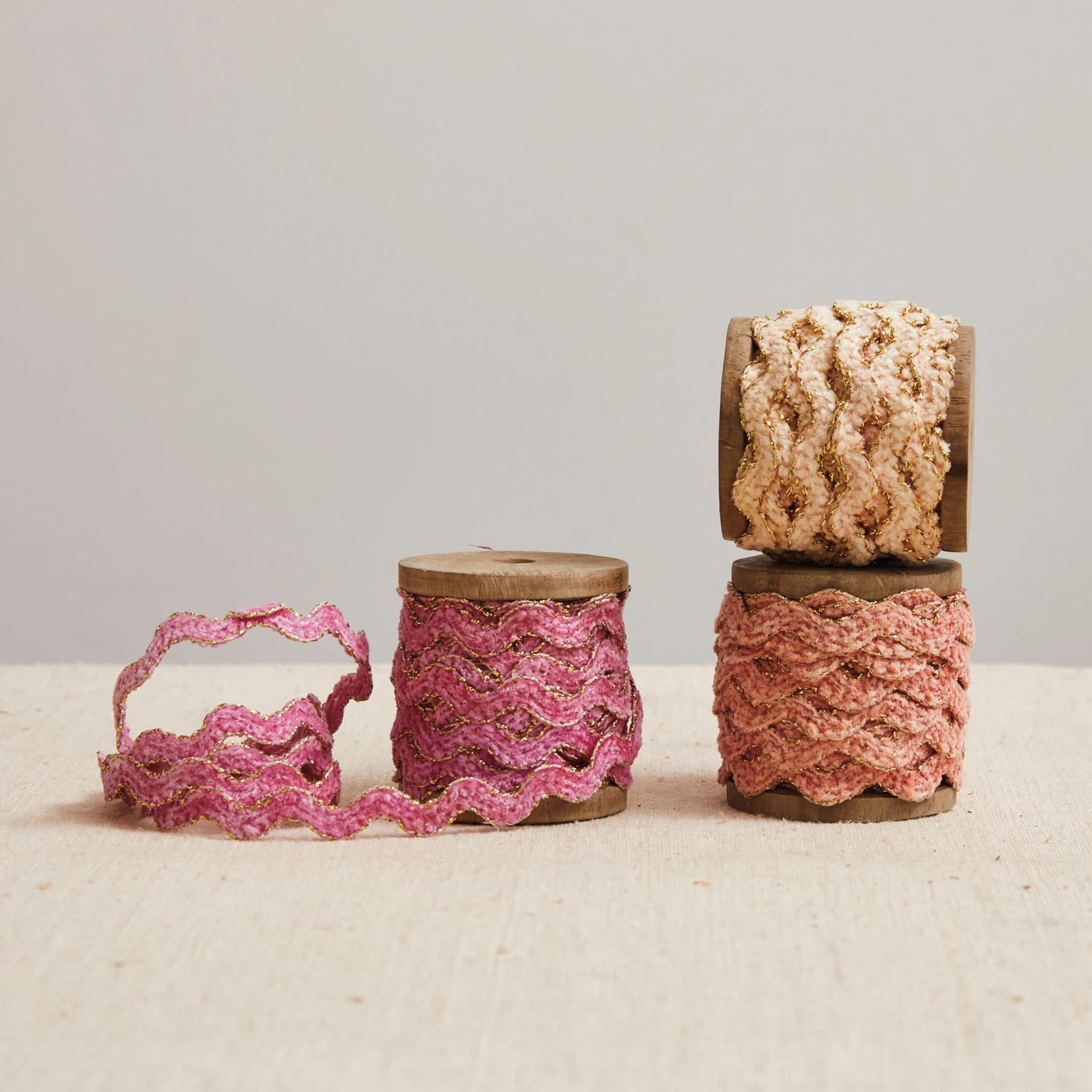The Creative Co-op Chenille Ribbon Spool Assorted features three wooden spools with pink, peach, and cream scalloped ribbons, perfect for gift wrapping. Displayed on a neutral fabric surface against a light background.