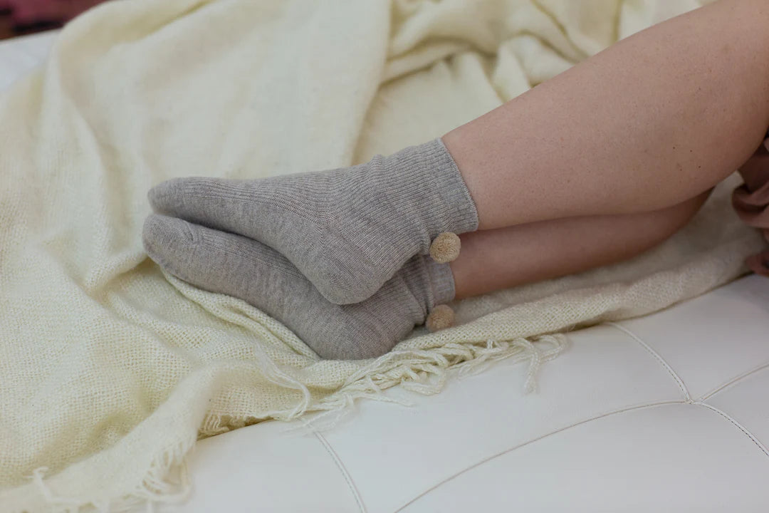 Someone wears Unified’s Winter Pom Pom Socks in cozy gray, resting their legs on a cream-colored blanket draped over a white surface.