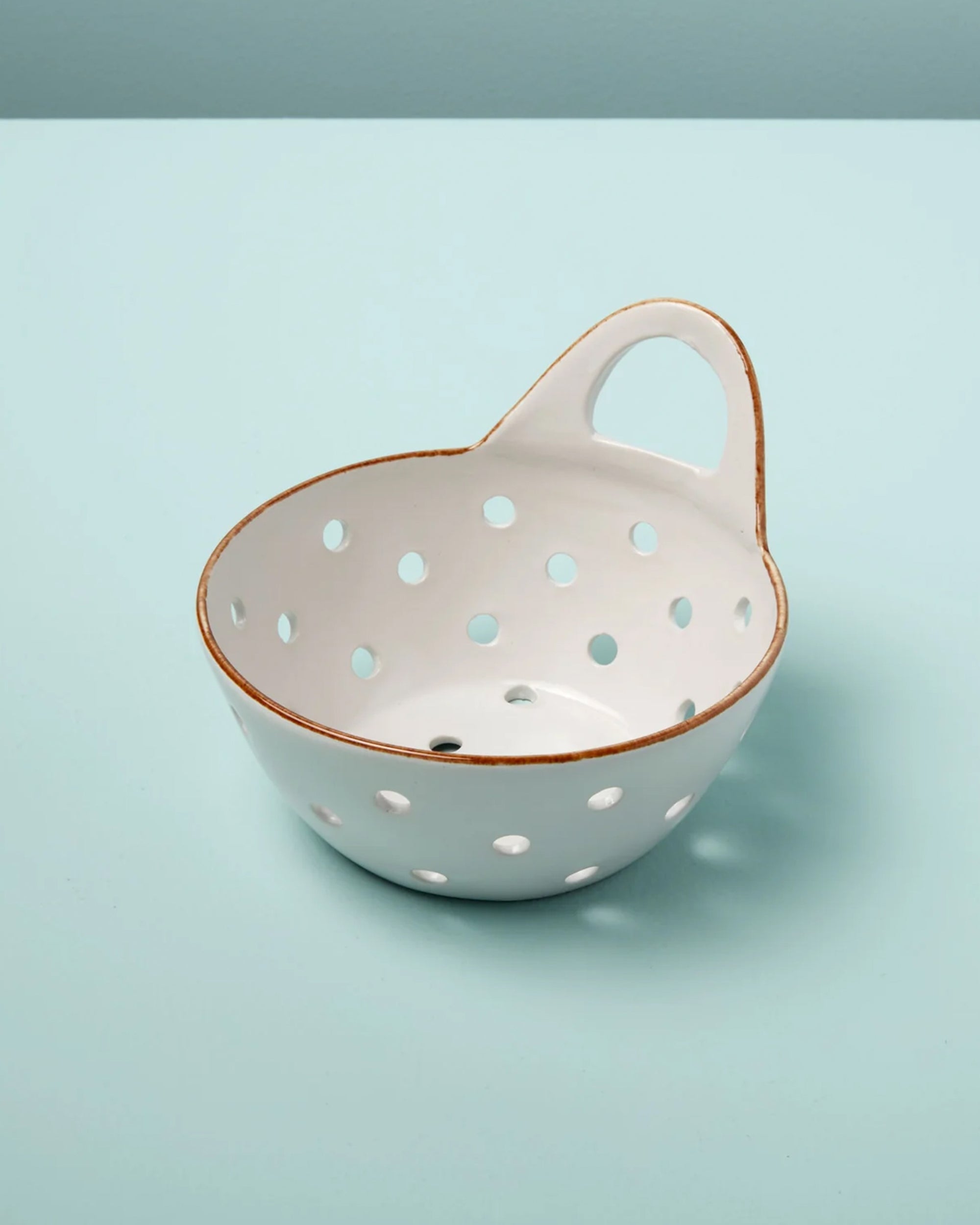 The Yara Colander Basket by Be Home is a handcrafted stoneware bowl featuring large circular holes, a single handle, and a rustic unglazed rim, shown resting on a light blue surface for added charm.